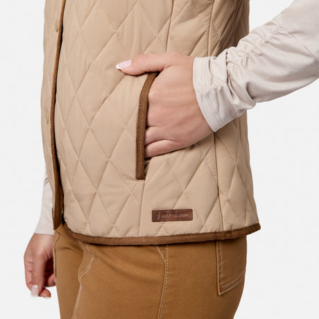 A person wearing the Free Country Women’s Cirrus Diamond Quilted Button Down Vest in beige and brown pants stands with one hand in their vest pocket. The lightweight insulated vest showcases diamond quilting and a small rectangular label near the hem.