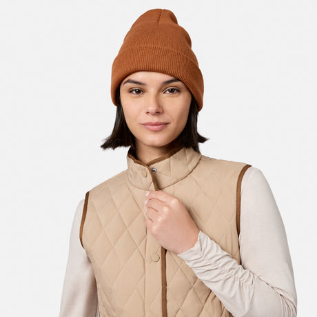 A woman with short dark hair, wearing a brown knit beanie, a long-sleeve light shirt, and the Free Country Women’s Cirrus Diamond Quilted Button Down Vest made from recycled polyester, stands against a plain white background.