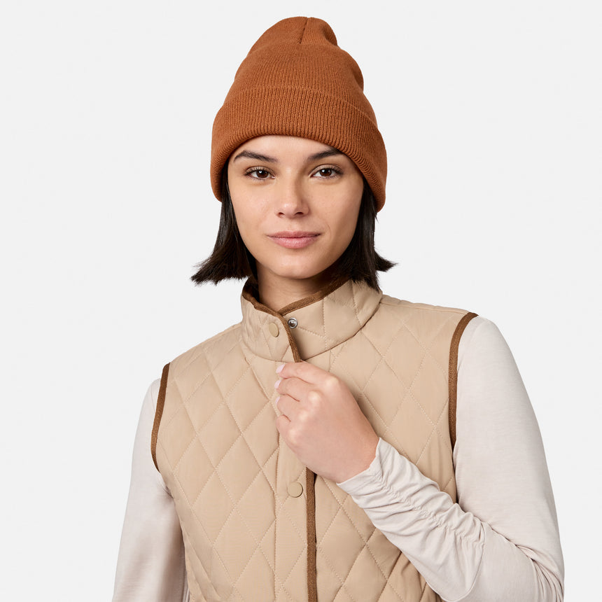 A woman with short dark hair, wearing a brown knit beanie, a long-sleeve light shirt, and the Free Country Women’s Cirrus Diamond Quilted Button Down Vest made from recycled polyester, stands against a plain white background.