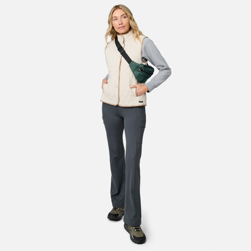 A woman wearing the Free Country Women’s Cirrus Diamond Quilted Button Down Vest in beige, paired with a gray long-sleeve top, dark gray pants, and black shoes, stands with one hand in her pocket and carries a green crossbody bag against a white background.