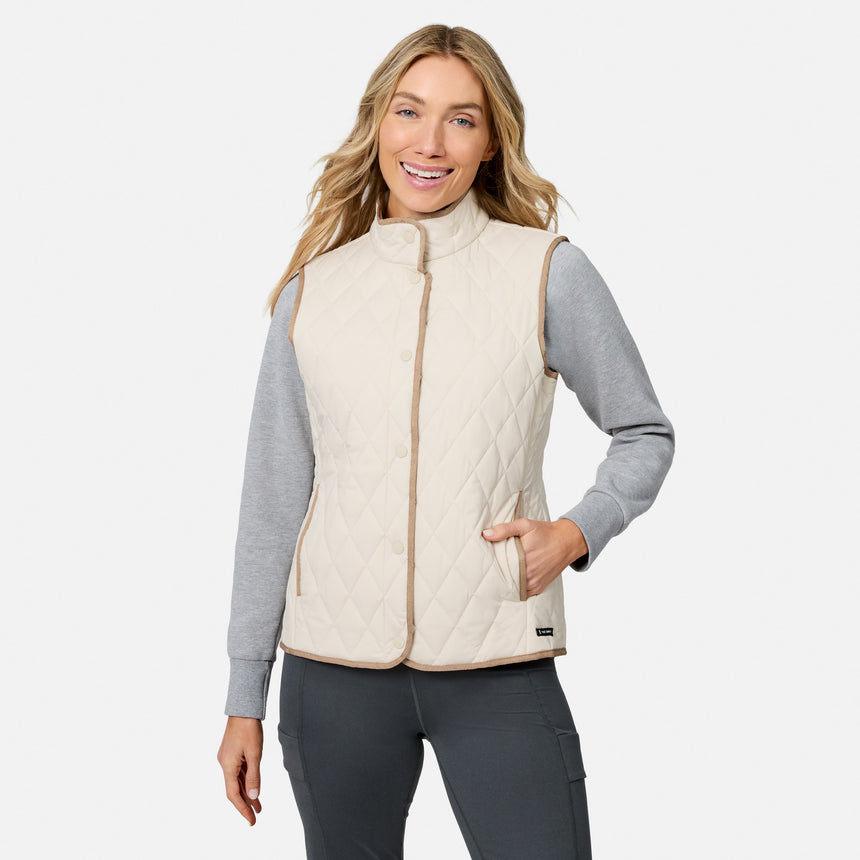 A smiling woman wears the Free Country Women’s Cirrus Diamond Quilted Button Down Vest over a gray long-sleeve shirt and dark pants, standing with one hand in her pocket against a plain white background.