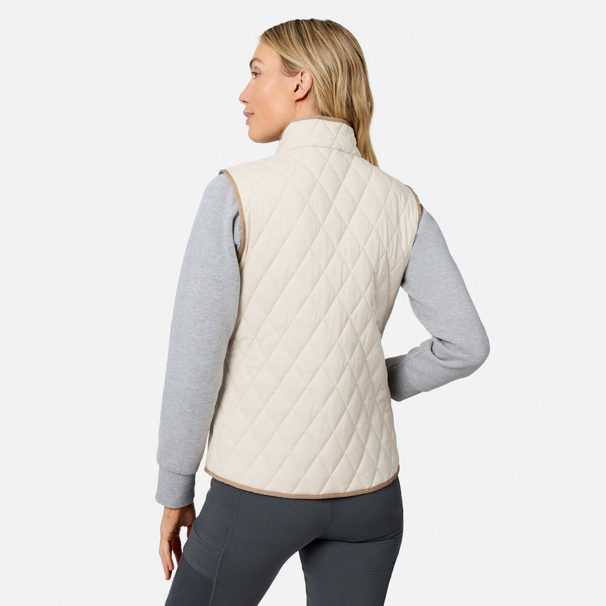 A woman facing away wears the Free Country Women’s Cirrus Diamond Quilted Button Down Vest in a light color over a gray long-sleeve shirt and dark pants, standing against a plain white background.