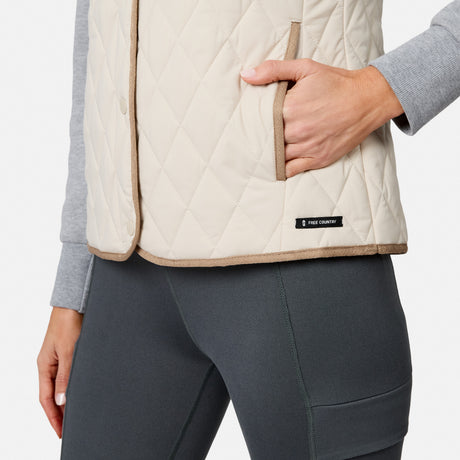 A person wearing the Free Country Women’s Cirrus Diamond Quilted Button Down Vest in beige, which features lightweight insulation, a pocket with one hand inside, and a small Free Country label near the hem, stands paired with gray leggings.