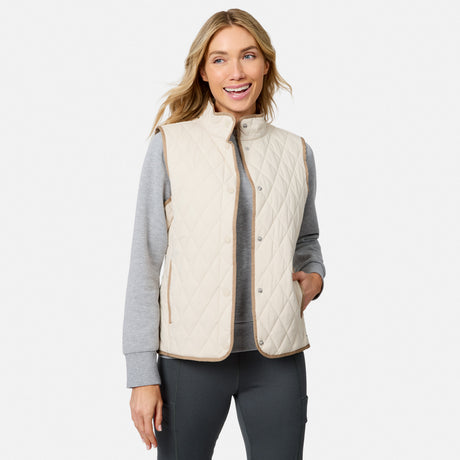 A woman wearing the Free Country Women’s Cirrus Diamond Quilted Button Down Vest in Bisque over a gray long-sleeve shirt and dark pants stands with one hand in her pocket, smiling against a white background.