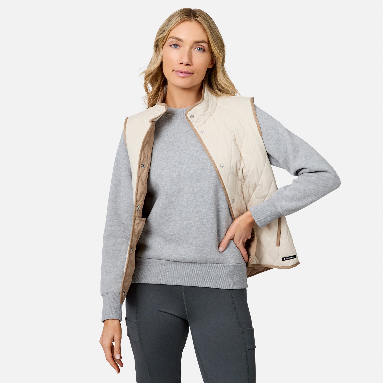 A woman stands against a plain white background, one hand in her pocket and a relaxed expression, wearing the Free Country Women’s Cirrus Diamond Quilted Button Down Vest in light beige over a gray sweatshirt and dark gray leggings.