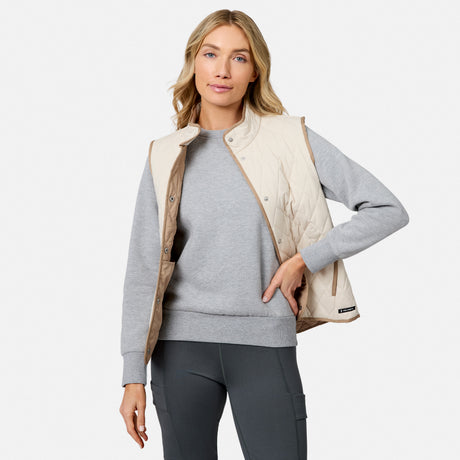 A woman stands against a plain white background, one hand in her pocket and a relaxed expression, wearing the Free Country Women’s Cirrus Diamond Quilted Button Down Vest in light beige over a gray sweatshirt and dark gray leggings.