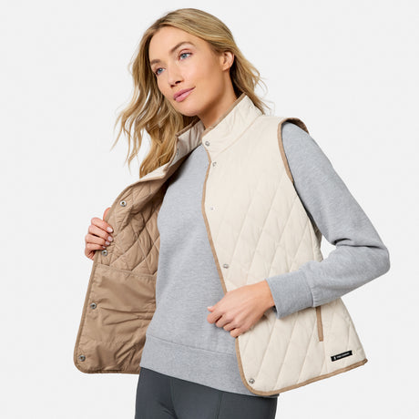A woman wearing a light gray sweatshirt and dark leggings opens the Free Country Women’s Cirrus Diamond Quilted Button Down Vest in beige, revealing its light brown interior. This stylish, lightweight insulated vest features a diamond quilted design that stands out against her plain, light background.