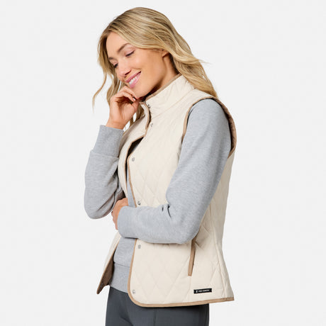 A woman wearing the Free Country Women’s Cirrus Diamond Quilted Button Down Vest over a gray sweatshirt smiles with her eyes closed, touching her face, against a plain white background.