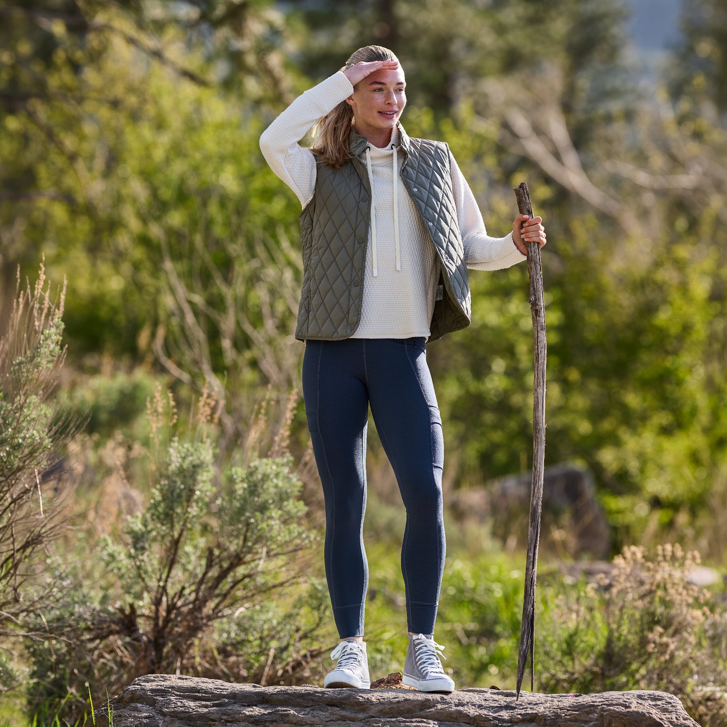A woman wearing the Free Country Women’s Cirrus Diamond Quilted Button Down Vest, along with a hoodie, leggings, and sneakers, stands outdoors on a log holding a walking stick. She is surrounded by greenery and trees, gazing into the distance with her hand shielding her eyes.