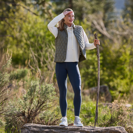 A woman wearing the Free Country Women’s Cirrus Diamond Quilted Button Down Vest, along with a hoodie, leggings, and sneakers, stands outdoors on a log holding a walking stick. She is surrounded by greenery and trees, gazing into the distance with her hand shielding her eyes.