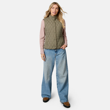 A woman stands against a plain white background wearing the Free Country Women’s Cirrus Diamond Quilted Button Down Vest in olive green, paired with a light pink long-sleeve shirt, wide-leg blue jeans, and brown shoes.