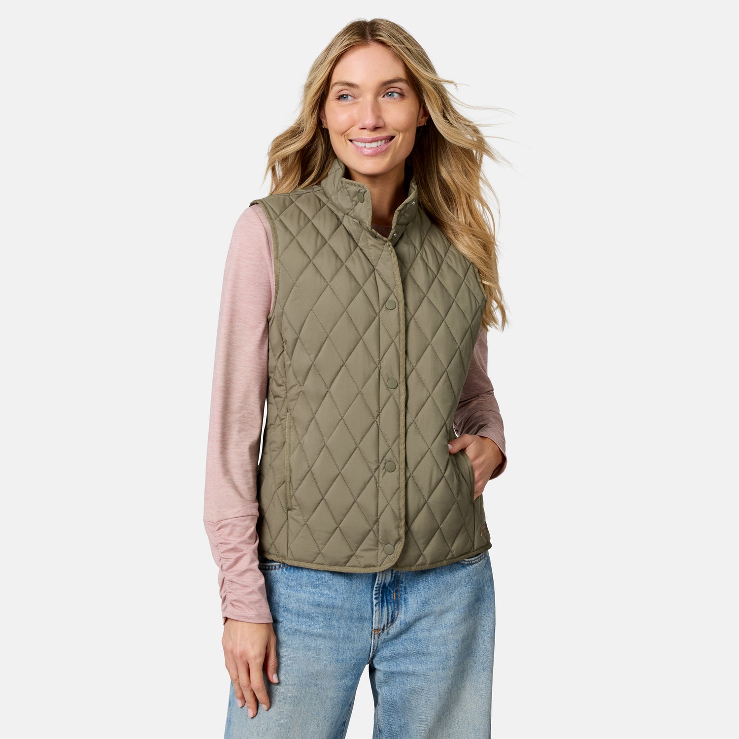 A woman stands smiling with one hand in her pocket against a plain background, wearing a light pink long-sleeve shirt, blue jeans, and the Free Country Women’s Cirrus Diamond Quilted Button Down Vest in olive green.