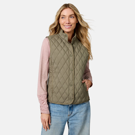 A woman stands smiling with one hand in her pocket against a plain background, wearing a light pink long-sleeve shirt, blue jeans, and the Free Country Women’s Cirrus Diamond Quilted Button Down Vest in olive green.