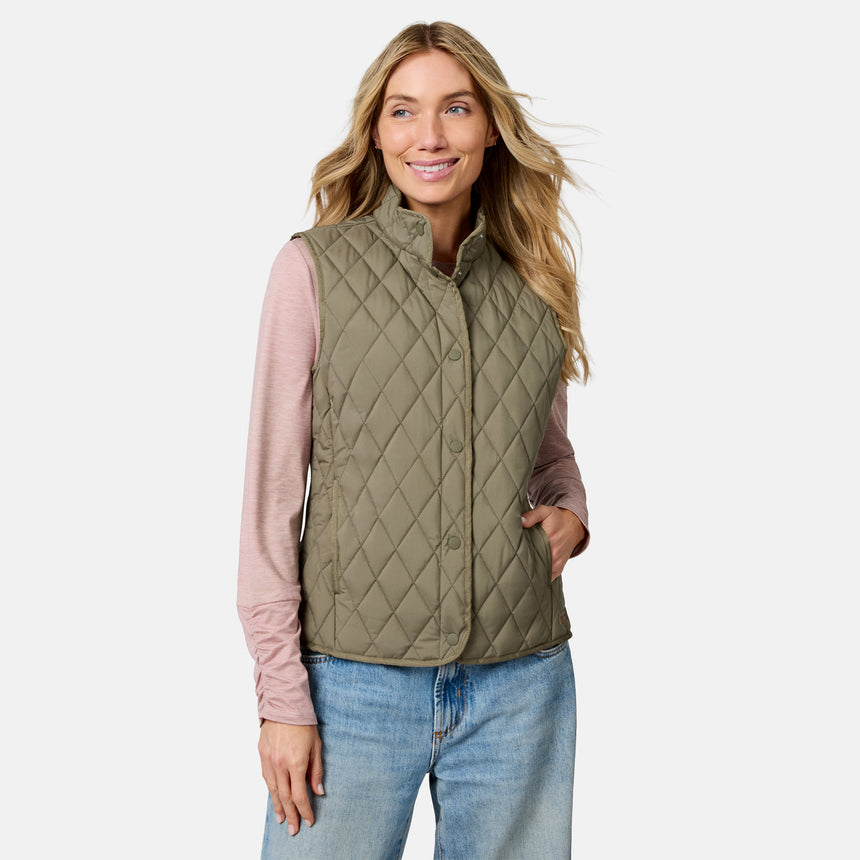 A woman stands smiling with one hand in her pocket against a plain background, wearing a light pink long-sleeve shirt, blue jeans, and the Free Country Women’s Cirrus Diamond Quilted Button Down Vest in olive green.