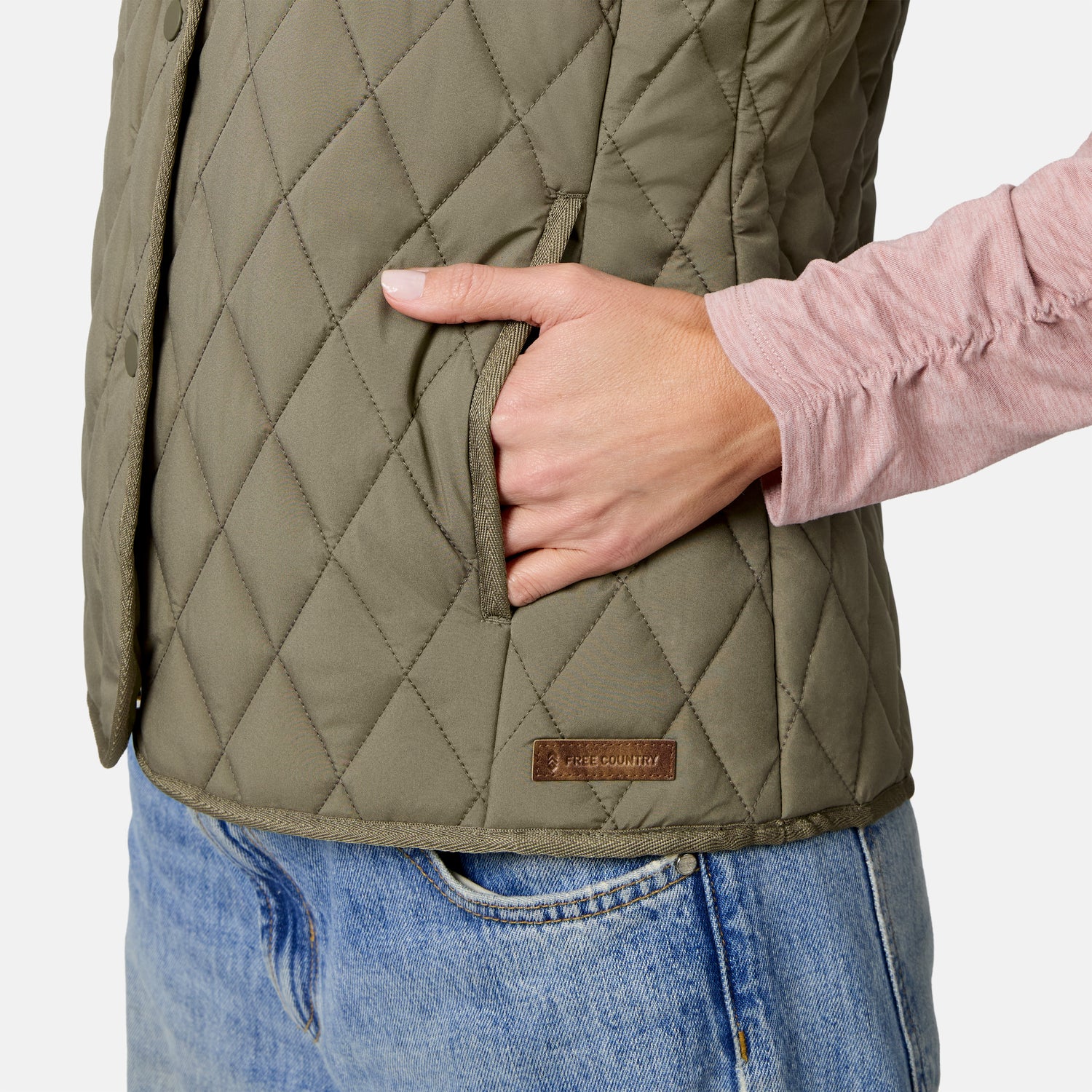 A person wearing the Free Country Women’s Cirrus Diamond Quilted Button Down Vest in green and blue jeans places their right hand in the vest pocket. They are also wearing a light pink long-sleeve shirt.