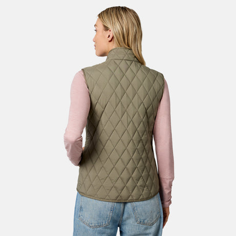 A woman with blonde hair is seen from the back against a plain white background, wearing the Free Country Women’s Cirrus Diamond Quilted Button Down Vest in olive green over a light pink long-sleeve shirt and blue jeans.