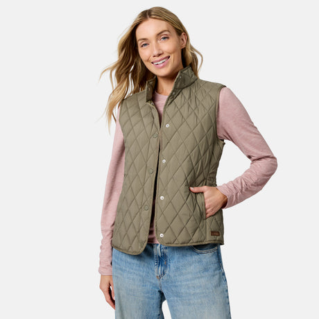 A smiling woman wears the Free Country Women’s Cirrus Diamond Quilted Button Down Vest in Olivine—crafted from recycled polyester—over a long-sleeve pink shirt and light blue jeans, with one hand in her vest pocket, standing against a plain light background.