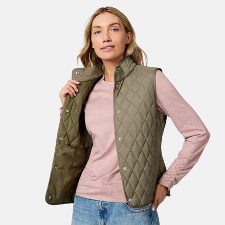 A woman in a pink long-sleeve shirt and blue jeans wears the Free Country Women’s Cirrus Diamond Quilted Button Down Vest in olive green, posing against a plain light background.