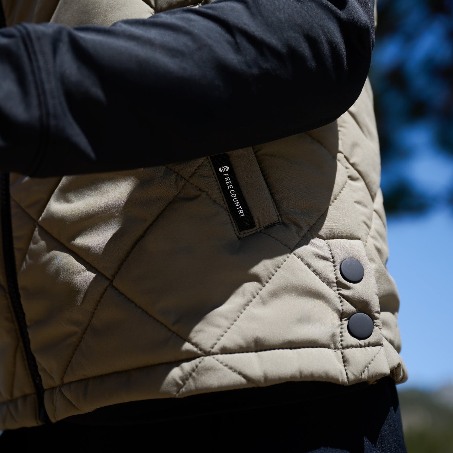 Close up of the hemline of a green quilted Free Country vest, with a Fee Country logo visible on the pocket