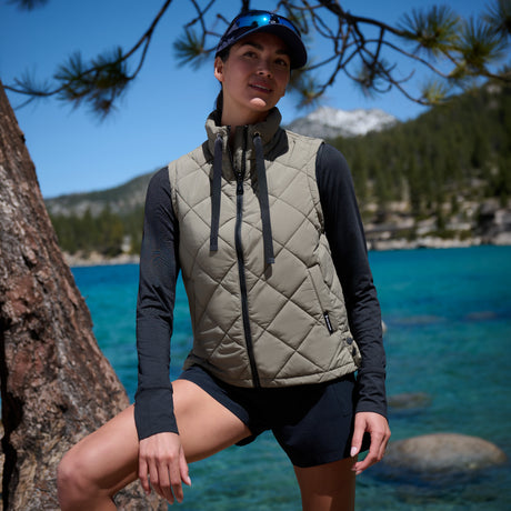 A woman outdoors by a turquoise lake wearing a quilted sleeveless vest over a dark long-sleeve top and shorts, with a tree trunk and forest in the background.