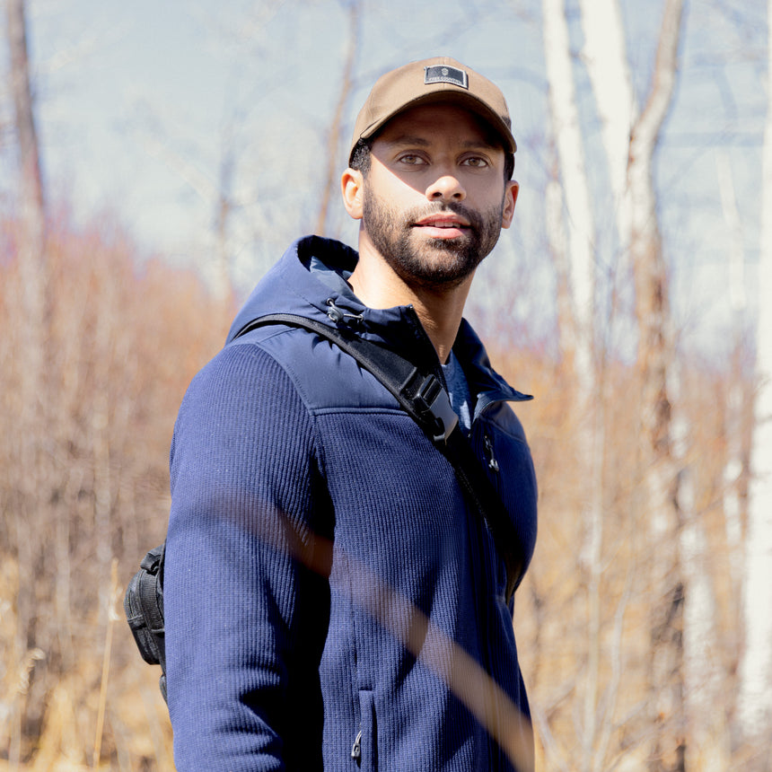 Man wearing a blue Free Country fleece jacket and brown cap outdoors with trees in the background