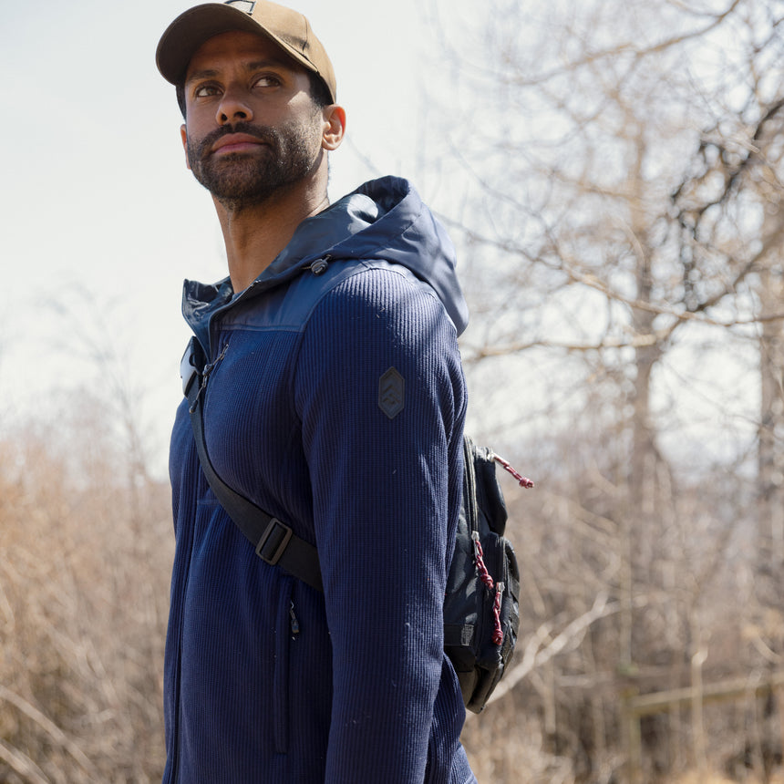 Man wearing a blue Free Country fleece jacket and brown cap outdoors with trees in the background
