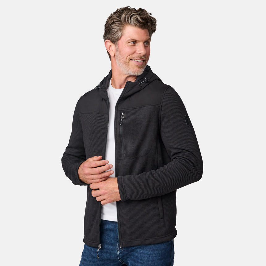 [Kolby is 6'2" wearing size M.] Men’s Free Country dark grey hooded jacket with full-zip front, zippered chest pocket, and dual side hand pockets—featuring textured fabric and a clean outdoor silhouette. Paired with beige pants for a rugged, cool-weather look.