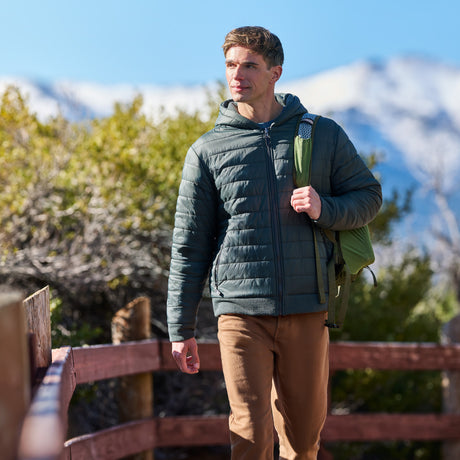 A man wearing a dark green quilted hooded puffer jacket, brown pants, and carrying a green backpack walks outdoors on a wooden path with mountains in the background.