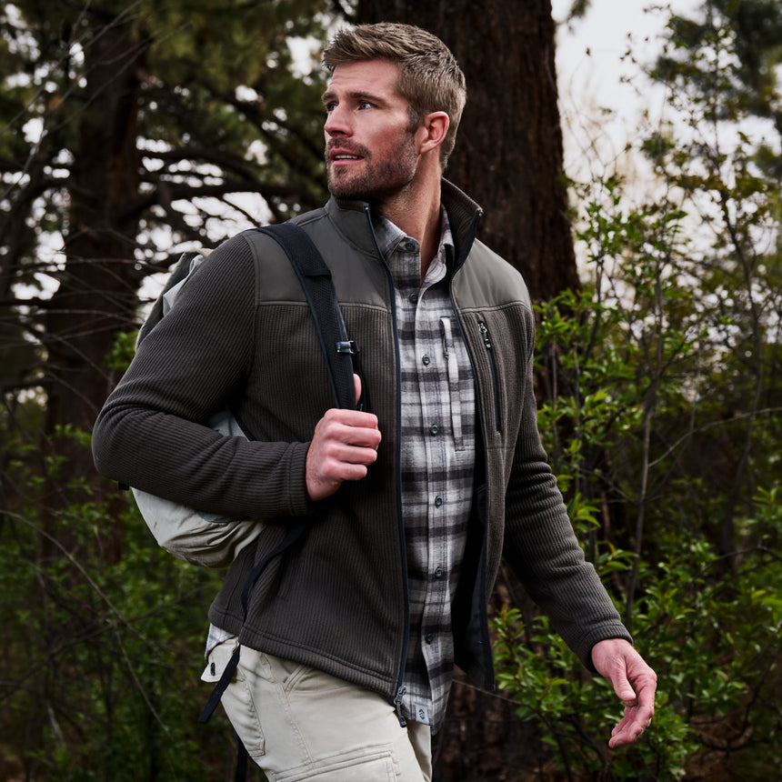 A man wearing a dark gray zip-up fleece jacket over a gray and white plaid flannel shirt, walking through a wooded outdoor setting.