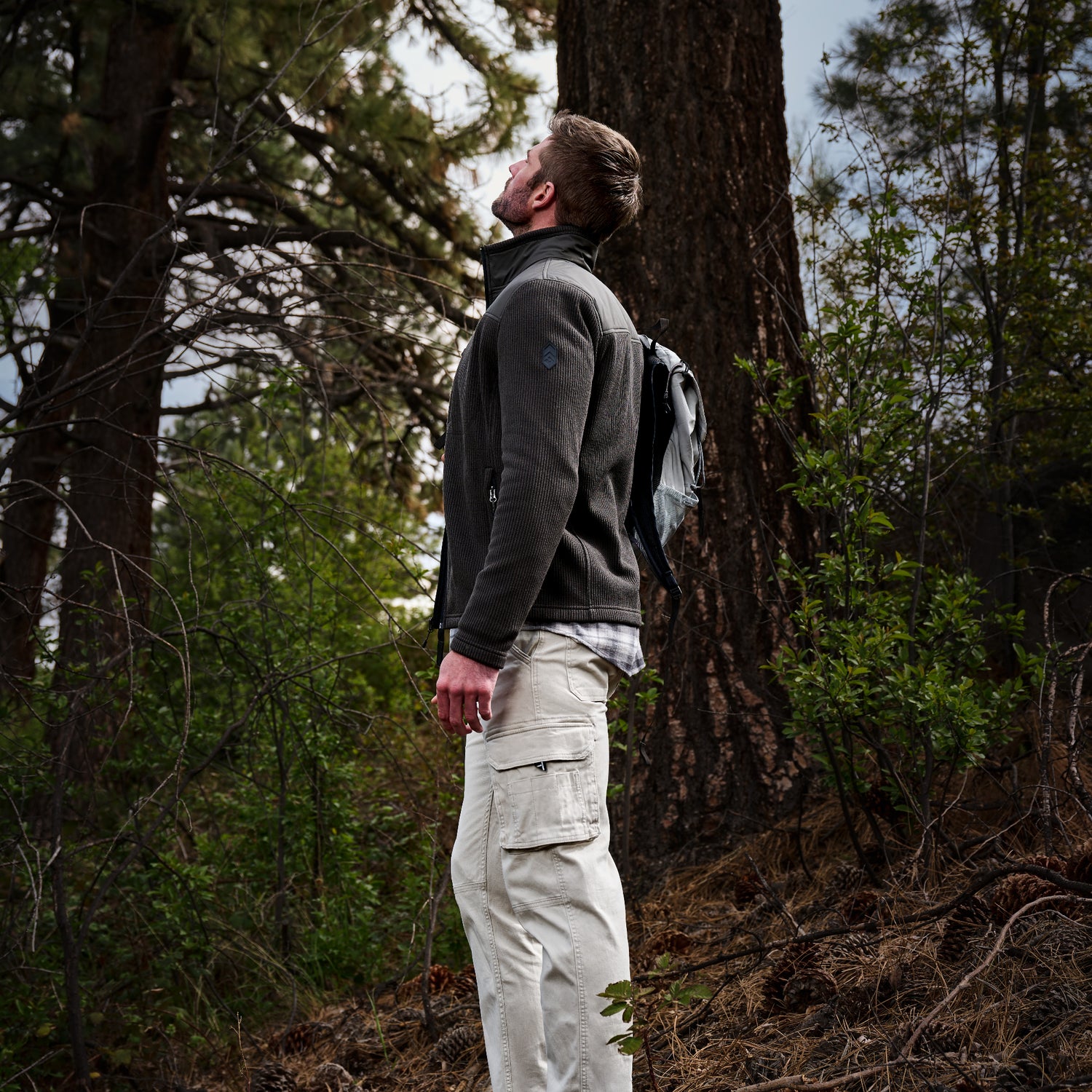 A man standing in a forest next to a large tree trunk, wearing a dark gray fleece jacket, light beige cargo pants, and a dark backpack.