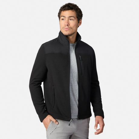 [Model is wearing size M.] A man wearing a black zip-up fleece jacket with a stand-up collar, ribbed shoulder panels, and zippered hand pockets, layered over a gray t-shirt and gray pants, against a light gray studio background.