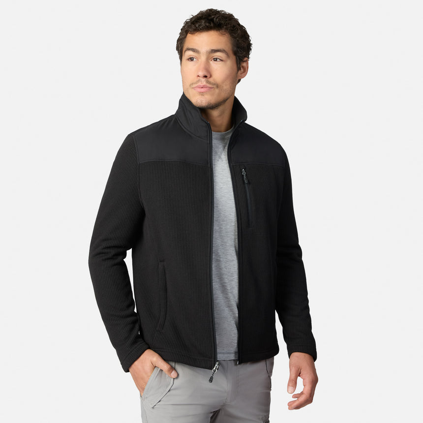[Model is wearing size M.] A man wearing a black zip-up fleece jacket with a stand-up collar, ribbed shoulder panels, and zippered hand pockets, layered over a gray t-shirt and gray pants, against a light gray studio background.