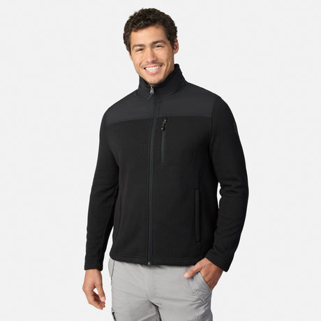 [Model is wearing size M.] A smiling man wearing a black long-sleeve zip-up fleece jacket with a stand-up collar and a vertical zippered chest pocket, paired with light gray pants, against a light gray studio background.