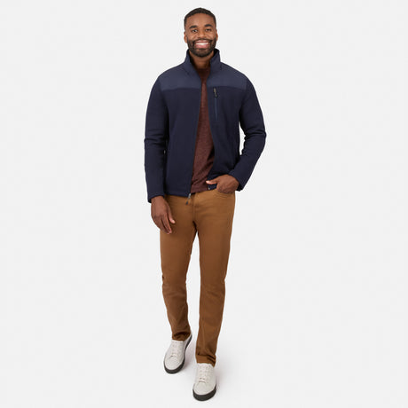 [Model is wearing size M.] A man modeling a navy blue zip-up fleece jacket with contrasting dark gray shoulder panels, worn over a brown shirt and paired with brown pants and white sneakers, against a light gray studio background.