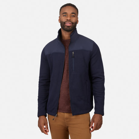 [Model is wearing size M.] A man wearing a navy blue zip-up fleece jacket with contrasting dark gray shoulder panels, layered over a brown sweater and paired with tan pants, against a light gray studio background.