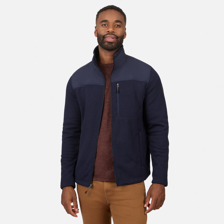 [Model is wearing size M.] A man wearing a navy blue zip-up fleece jacket with contrasting dark gray shoulder panels, layered over a brown sweater and paired with tan pants, against a light gray studio background.