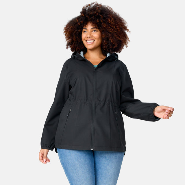 A woman with curly hair is wearing the Free Country Women's Plus Size X2O Anorak Rain Jacket in black and blue jeans, smiling and posing against a plain light gray background.