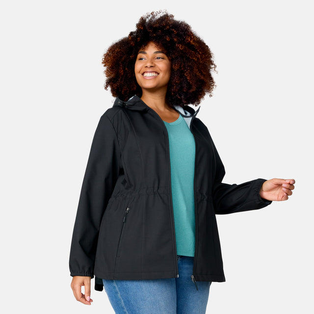 A woman with curly hair is smiling and standing against a plain light background, wearing the Free Country Women's Plus Size X2O Anorak Rain Jacket over a teal top and blue jeans.