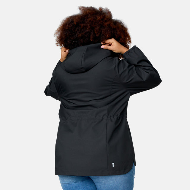A woman with curly hair, dressed in the Free Country Women's Plus Size X2O Anorak Rain Jacket in black and blue jeans, faces away from the camera while adjusting the hood. The background is plain white.