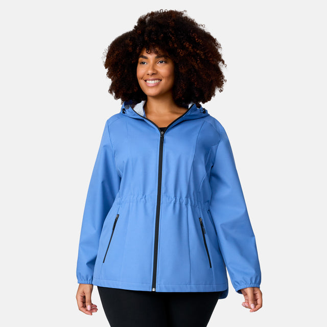 A woman with curly hair smiles in a Free Country Women's Plus Size X2O Anorak Rain Jacket in light blue, featuring black zipper details, paired with black pants, against a plain light gray background.