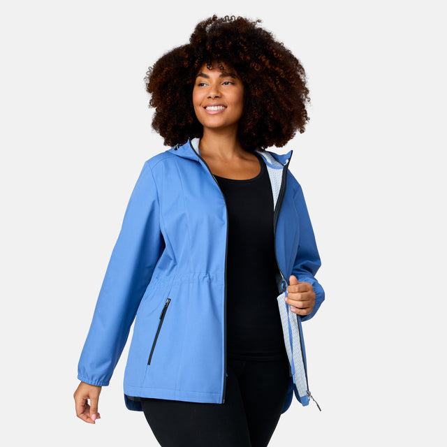 A woman with curly hair smiles against a light background, wearing the Free Country Women's Plus Size X2O Anorak Rain Jacket in light blue over a black top and pants, holding it open to highlight its stylish, practical water-resistant design.