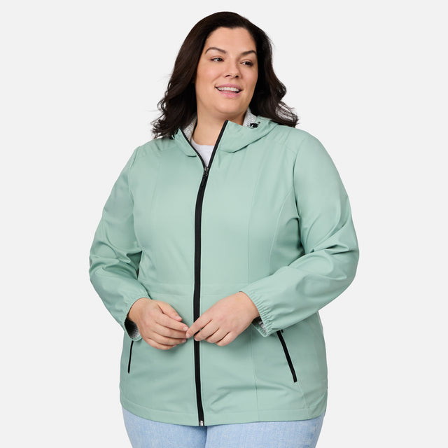 A woman with dark hair gently smiles, standing against a white background in the Free Country Women's Plus Size X2O Anorak Rain Jacket—a light green, water-resistant hooded jacket—with her hands loosely clasped in front.