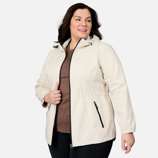 A woman with long brown hair wears the Women's Plus Size X2O Anorak Rain Jacket by Free Country in cream over a brown shirt and black pants, standing against a plain light background and smiling as she looks to the side.