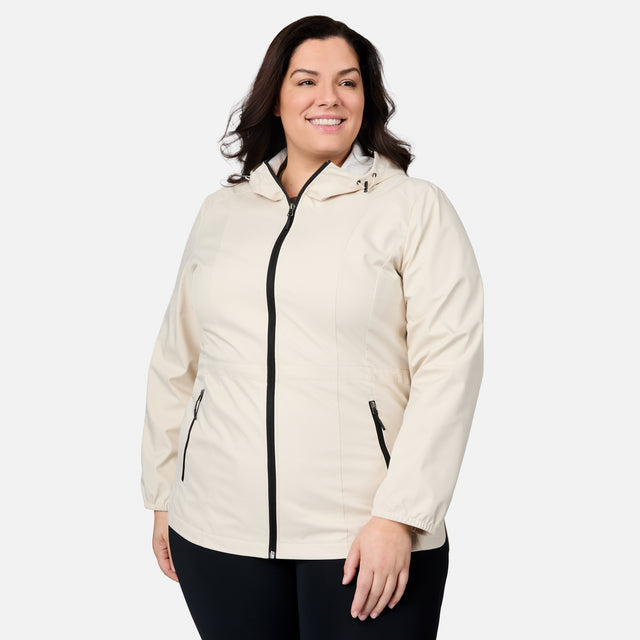 A woman wearing the Women's Plus Size X2O Anorak Rain Jacket by Free Country, featuring a light beige water-resistant design with black zippers, stands against a plain white background, smiling and looking slightly to the side.