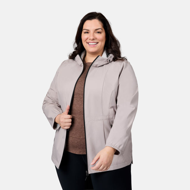 A smiling woman with long dark hair models the Free Country Women's Plus Size X2O Anorak Rain Jacket in light gray over a brown top and black pants, posing with one hand on the zipper against a plain white background.