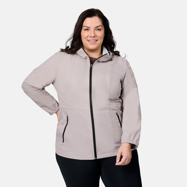 A woman with long dark hair smiles while wearing the Free Country Women's Plus Size X2O Anorak Rain Jacket in light gray with black zipper details, paired with black pants and standing against a plain white background.