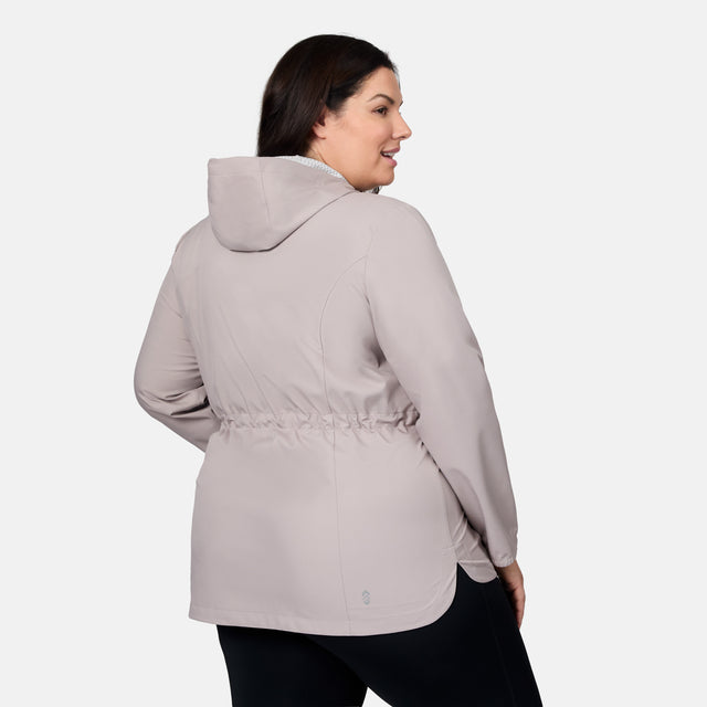 A woman models the Free Country Women's Plus Size X2O Anorak Rain Jacket in light gray, featuring a cinched waist and long sleeves. She pairs the water-resistant jacket with black pants against a plain white background.