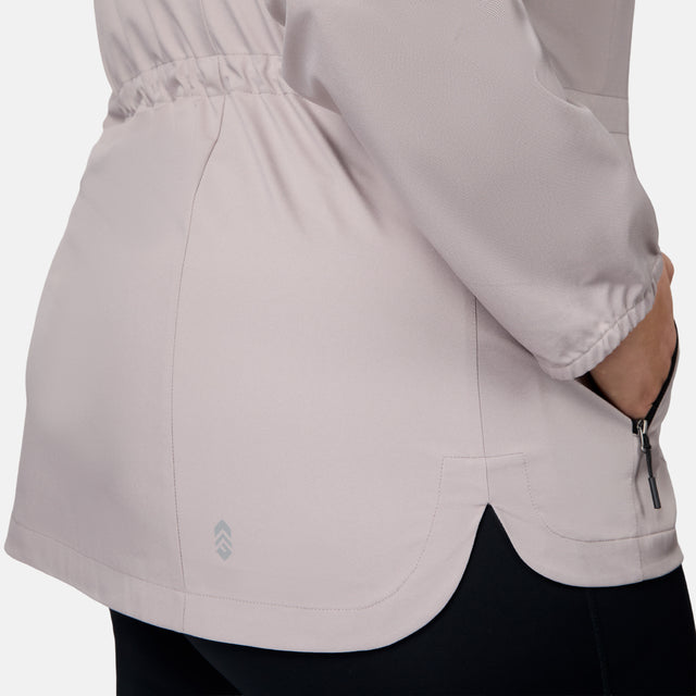 A close-up shows a person wearing the Free Country Women's Plus Size X2O Anorak Rain Jacket, featuring a light color, water-resistant fabric, elastic waistband, and zippered side pocket. The hand is partly in the pocket, with black pants visible underneath.