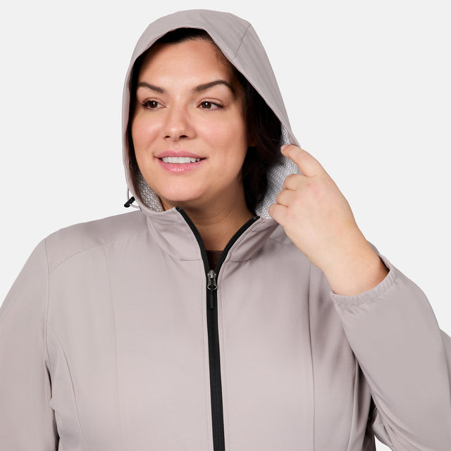 A woman wears the Free Country Women's Plus Size X2O Anorak Rain Jacket in light gray with a hood, smiling and holding the hood's edge against a plain white background.
