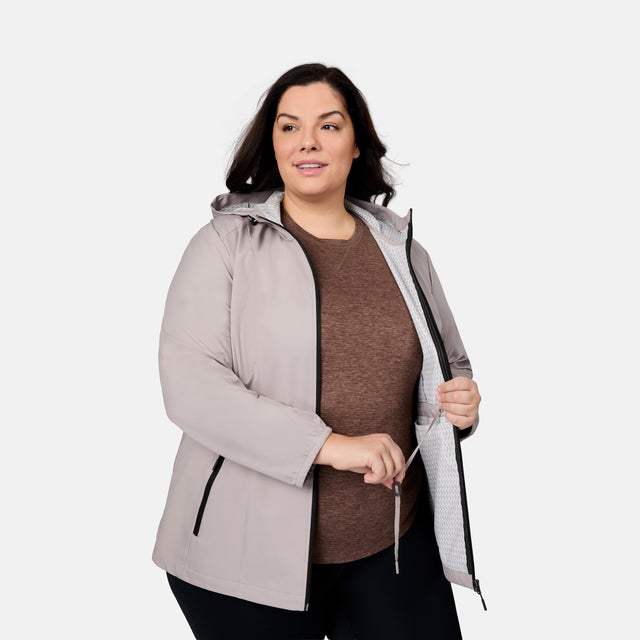 A woman wearing the Free Country Women's Plus Size X2O Anorak Rain Jacket in light gray over a brown shirt stands against a white background, holding the partially unzipped water-resistant jacket open with one hand.